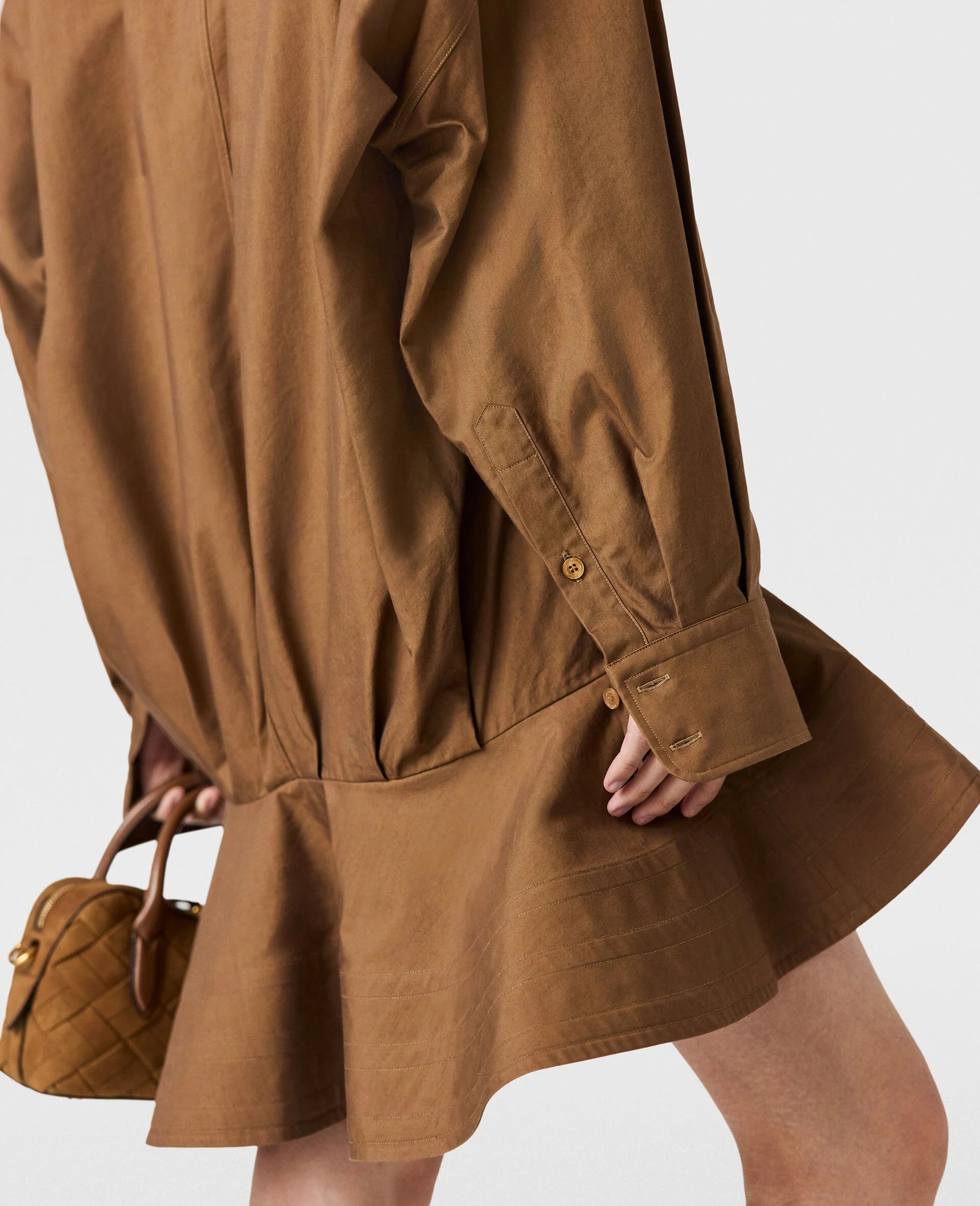 A person holds a small quilted brown handbag, wearing a Stella McCartney Washed Cotton Peplum Shirt Dress. Only the lower half of the dress and their hand are visible.