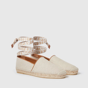The Gaia Canvas Flat by Stella McCartney is a pair of beige espadrilles made from lightweight, textured canvas with woven soles and ankle straps adorned with the Stella McCartney logo in white lettering.