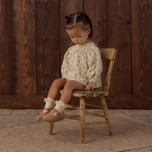 A toddler with pigtails sits on an old wooden chair, dressed in the Rylee & Cru Baby Girls’ Quincy Set—a coordinated long-sleeve top and matching bloomer—paired with ruffled socks and brown shoes, eyes closed and facing down against a wooden wall backdrop.