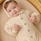 A baby lies in a woven basket, wearing the Quincy Mae Baby Girls' Long Sleeve Bubble Romper in cream, featuring a hood and crafted from cozy pointelle fabric.