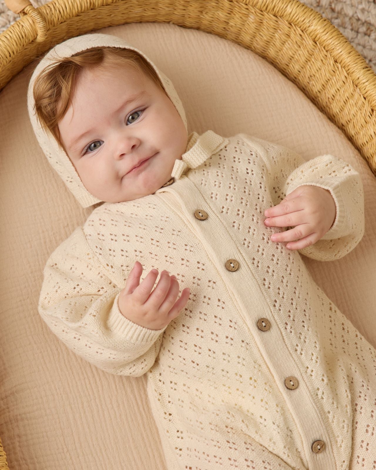 A baby lies in a woven basket, wearing the Quincy Mae Baby Girls' Long Sleeve Bubble Romper in cream, featuring a hood and crafted from cozy pointelle fabric.