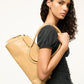 A person with long curly hair holds a beige Staud Maude Shoulder Bag in polished leather. Wearing a black top and blue jeans, she stands facing forward in a neutral pose, the bag's flap closure elegantly complementing her look.