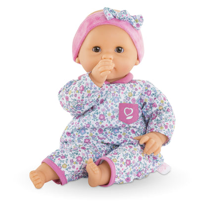 Corolle’s Bébé Calin Capucine baby doll, dressed in floral pajamas and a pink headband with a bow and crafted from soft vinyl, sits sweetly with one thumb in its mouth while the other hand rests on its leg.
