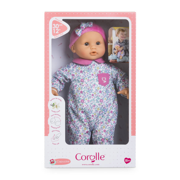 The Corolle Bébé Calin Capucine is a 12-inch baby doll dressed in a floral outfit and pink hat, crafted from soft vinyl, and presented in a pink and white branded box featuring a photo of a child holding the doll.