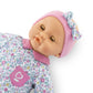 The Corolle Bébé Calin Capucine baby doll is dressed in a floral-patterned outfit with a pink collar and pocket, complemented by a pink headband with a bow. Crafted from soft vinyl, she is shown against a white background.