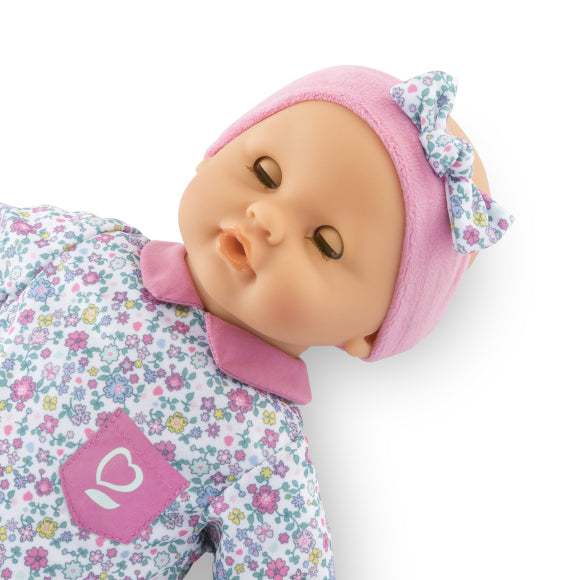 The Corolle Bébé Calin Capucine baby doll is dressed in a floral-patterned outfit with a pink collar and pocket, complemented by a pink headband with a bow. Crafted from soft vinyl, she is shown against a white background.