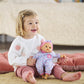A young child sits on the floor, smiling and holding a Corolle Bébé Calin Capucine doll, which is made of soft vinyl and dressed in a floral outfit with a pink hat, with a bed in the background.