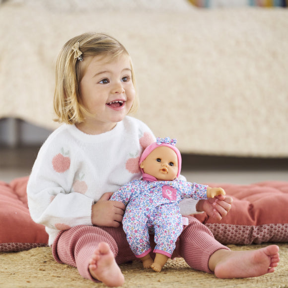 A young child sits on the floor, smiling and holding a Corolle Bébé Calin Capucine doll, which is made of soft vinyl and dressed in a floral outfit with a pink hat, with a bed in the background.