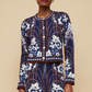 A woman wearing the Cara Cara Livi Jacket, a blue jacquard knit cardigan with white and brown floral designs, stands against a beige background.