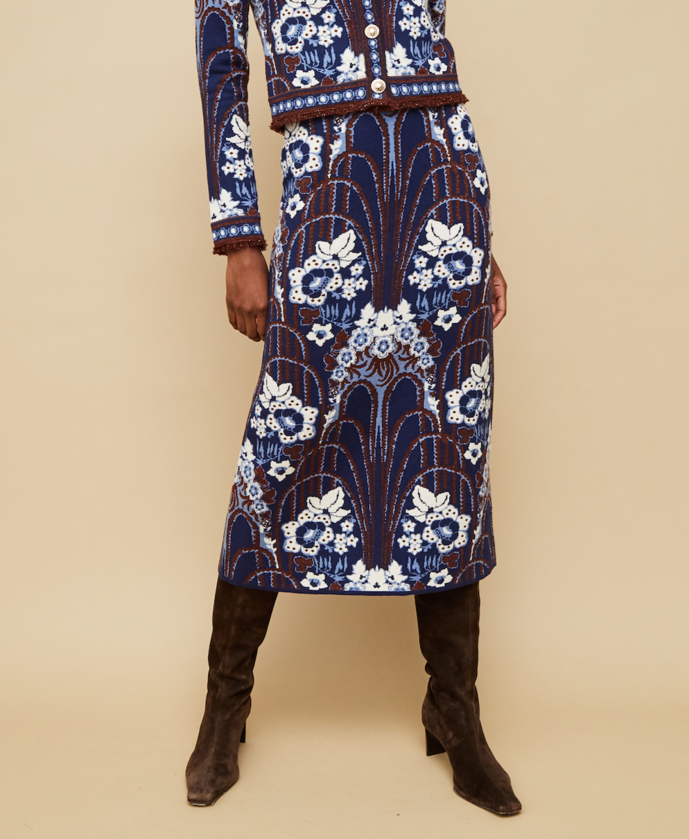A person models the Cara Cara Clara Skirt and matching blue jacquard jacket with white floral patterns, paired with dark brown knee-high boots, against a beige background.