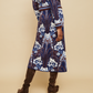 A person wearing the Clara Skirt by Cara Cara, featuring a blue jacquard pattern with white floral designs and dark brown knee-high boots, stands against a beige background.