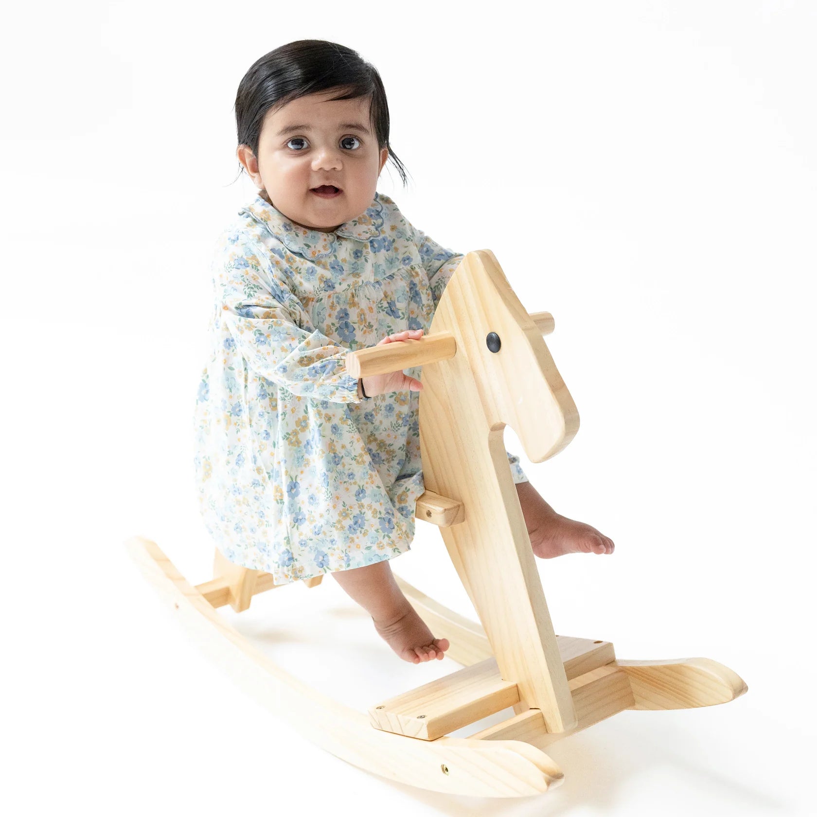 A baby wearing the Angel Dear Baby Girls' Long Sleeve Petal Collar Dress and Bloomer sits on a light wooden rocking horse against a white background.