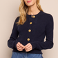 A woman wears the Cara Cara Ellery Cardigan—navy blue, long-sleeve, crewneck with gold floral buttons—paired with blue jeans, standing against a beige background.