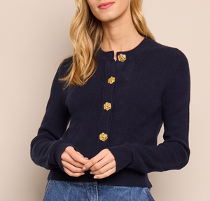 A woman wears the Cara Cara Ellery Cardigan—navy blue, long-sleeve, crewneck with gold floral buttons—paired with blue jeans, standing against a beige background.