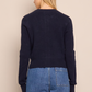A woman faces away from the camera, wearing the Cara Cara Ellery Cardigan—navy blue with gold floral buttons—and blue jeans, set against a beige background.