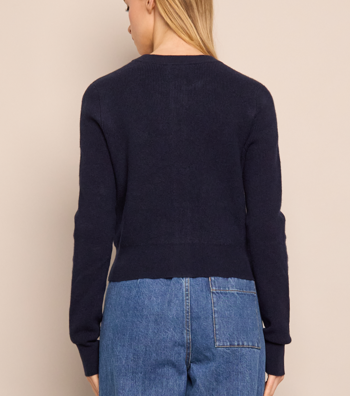 A woman faces away from the camera, wearing the Cara Cara Ellery Cardigan—navy blue with gold floral buttons—and blue jeans, set against a beige background.