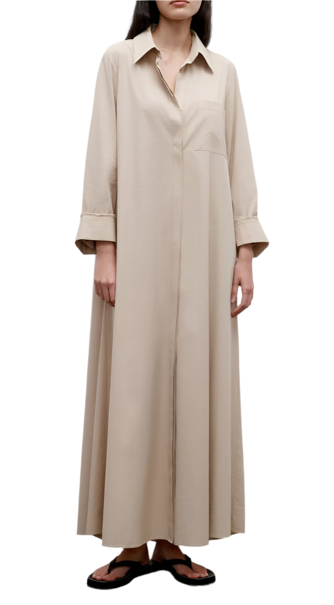 A woman wears the Jenny's Crystal Gown by TWP, an oversized maxi shirt dress in beige Italian cotton shirting with a collar, chest pocket, and rolled-up sleeves, paired with black sandals.