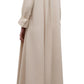A person in TWP's Jenny's Crystal Gown, a luxurious maxi dress, stands sideways and faces away, pairing the look with black sandals and a long beige coat.