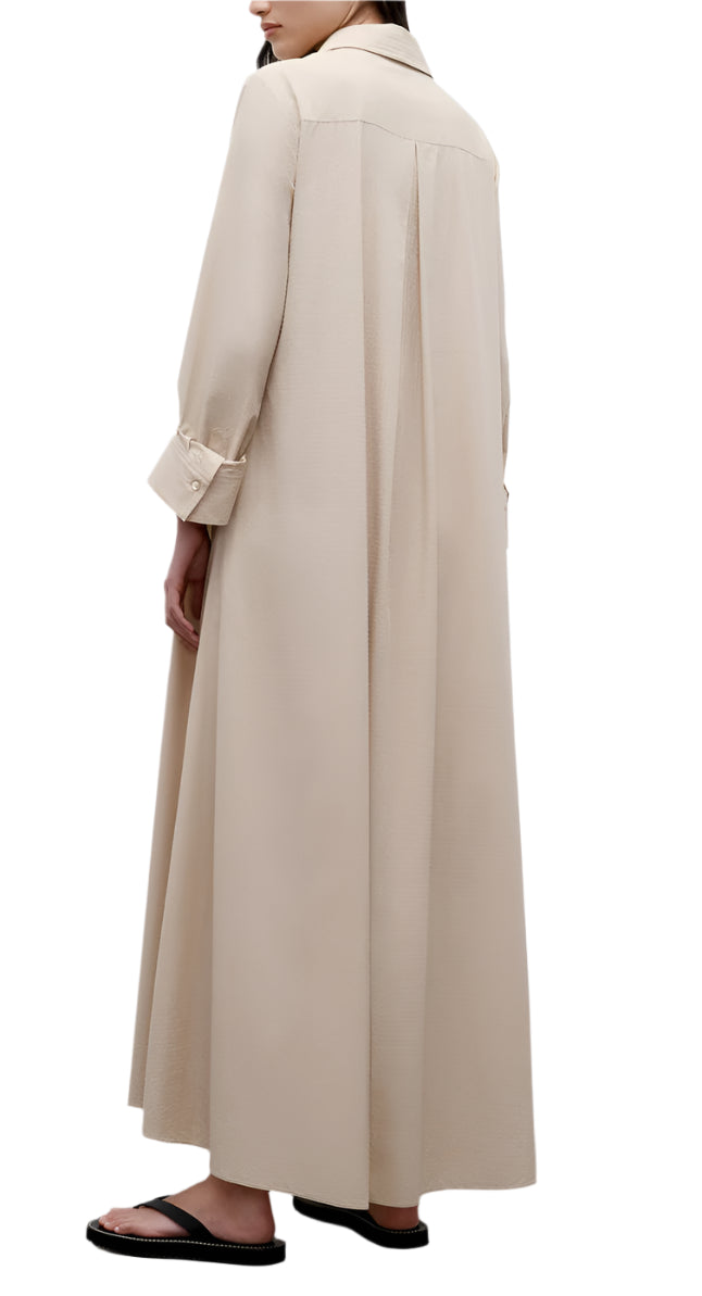 A person in TWP's Jenny's Crystal Gown, a luxurious maxi dress, stands sideways and faces away, pairing the look with black sandals and a long beige coat.
