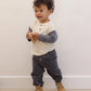 A toddler with curly hair stands against a plain wall, wearing a long-sleeve shirt, tan boots, and the Quincy Mae Baby Boys’ and Boys’ Luca Pant—gray corduroy pants with an elastic waistband—while holding a wooden toy.