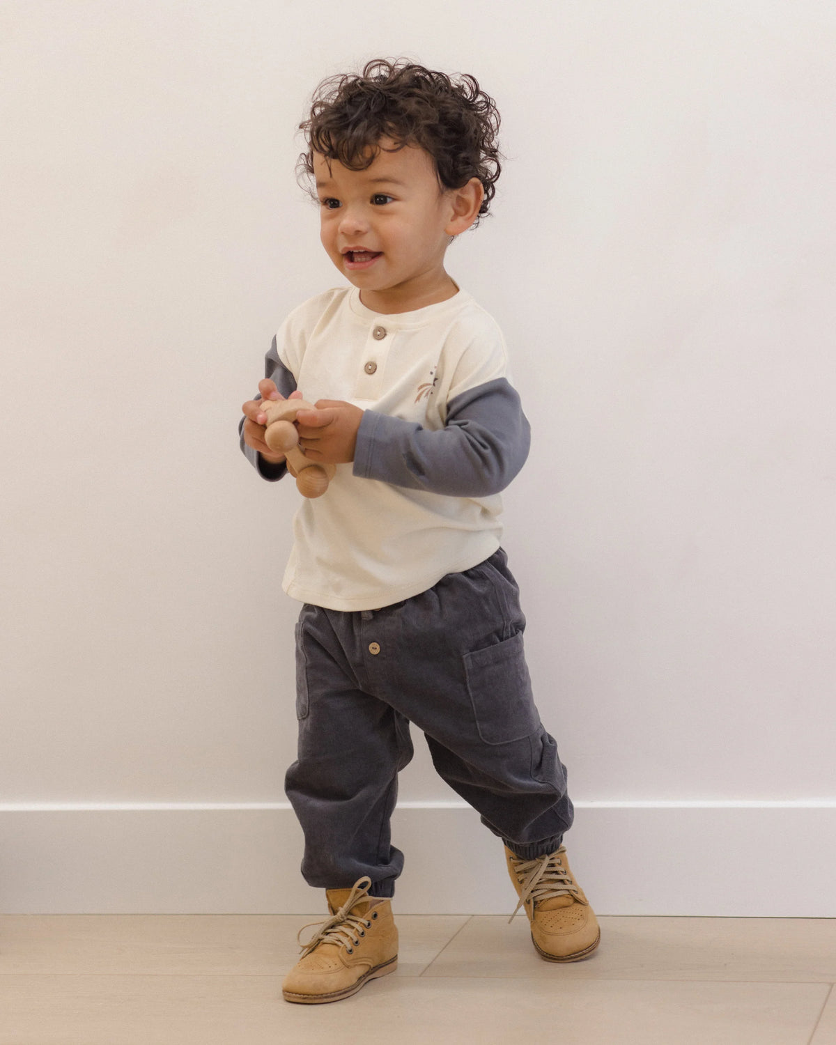 A toddler with curly hair stands against a plain wall, wearing a long-sleeve shirt, tan boots, and the Quincy Mae Baby Boys’ and Boys’ Luca Pant—gray corduroy pants with an elastic waistband—while holding a wooden toy.