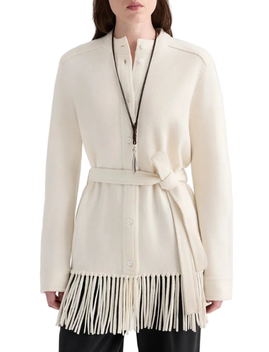 A person wears the TWP Thea Italian Cashmere Jacket, a cream-colored belted knit style featuring fringe trim at the hem and buttoned front, paired with a long necklace.