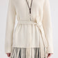 A person wearing TWP’s Thea Italian Cashmere Jacket—a cream knit style with a tie belt and elegant fringe trim at the hem—paired with a long necklace featuring a leaf-shaped pendant.