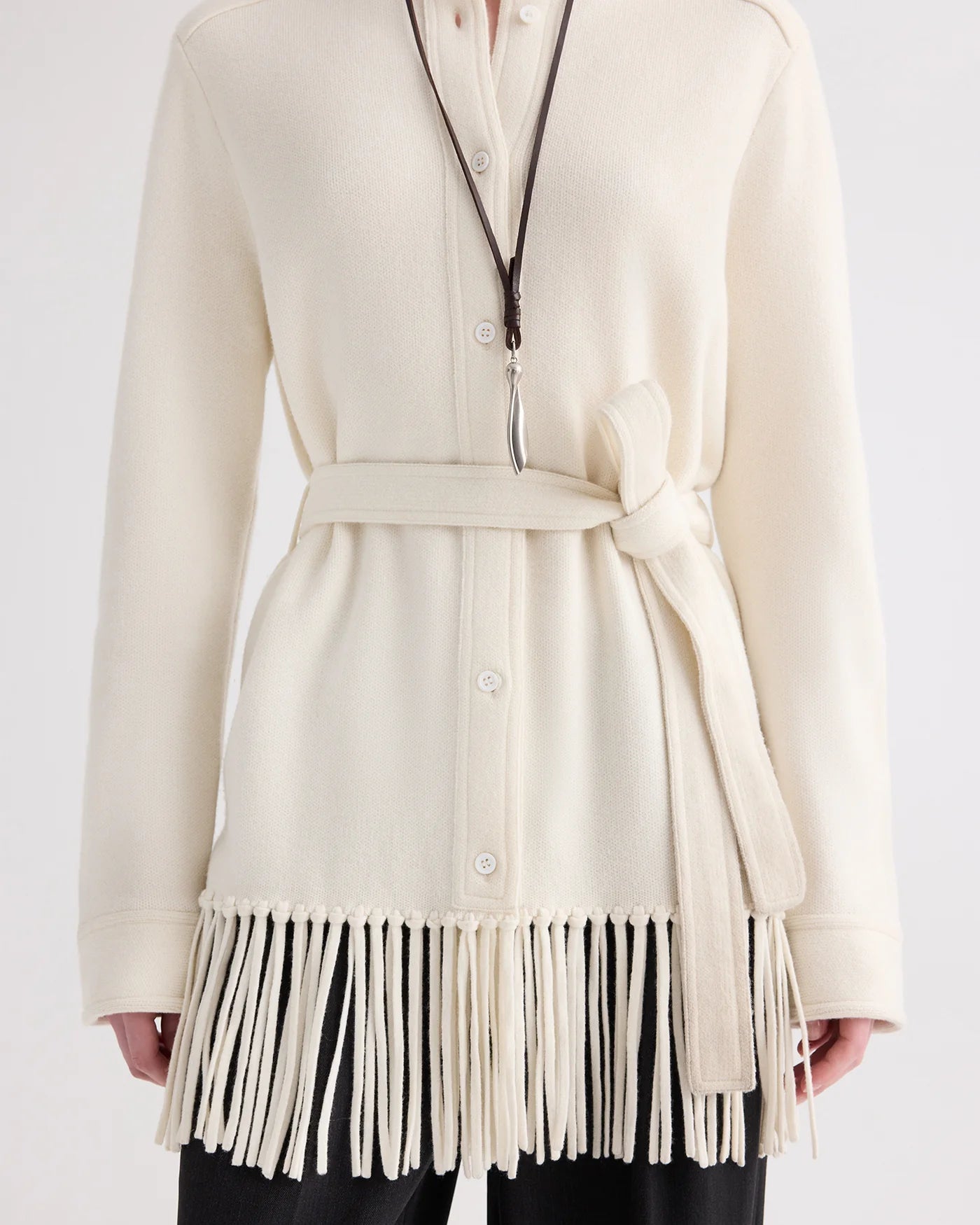 A person wearing TWP’s Thea Italian Cashmere Jacket—a cream knit style with a tie belt and elegant fringe trim at the hem—paired with a long necklace featuring a leaf-shaped pendant.