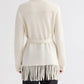 A person is seen from the back against a plain background, wearing the TWP Thea Italian Cashmere Jacket—an off-white piece featuring a belted waist and fringe trim along the hem.