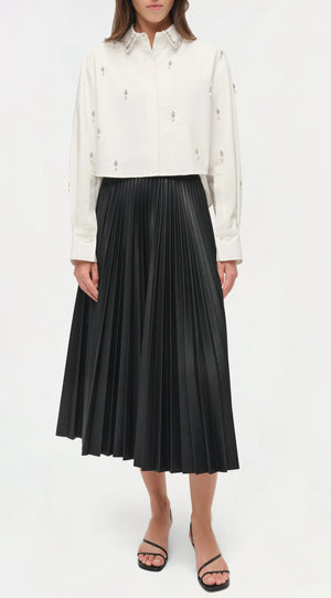 A person wears the Simkhai Renata Embellished Cropped Shirt, a white long-sleeve blouse with crystal pearl details, paired with a black pleated midi skirt and black sandals, standing against a plain white background.