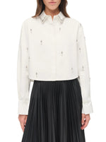 A woman wears the Simkhai Renata Embellished Cropped Shirt, a white cotton poplin top with jeweled details, styled with a black pleated skirt.