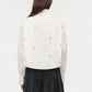 A person with long brown hair is seen from the back wearing the Simkhai Renata Embellished Cropped Shirt—white cotton poplin with crystal pearl details—and a black pleated skirt, set against a plain background.