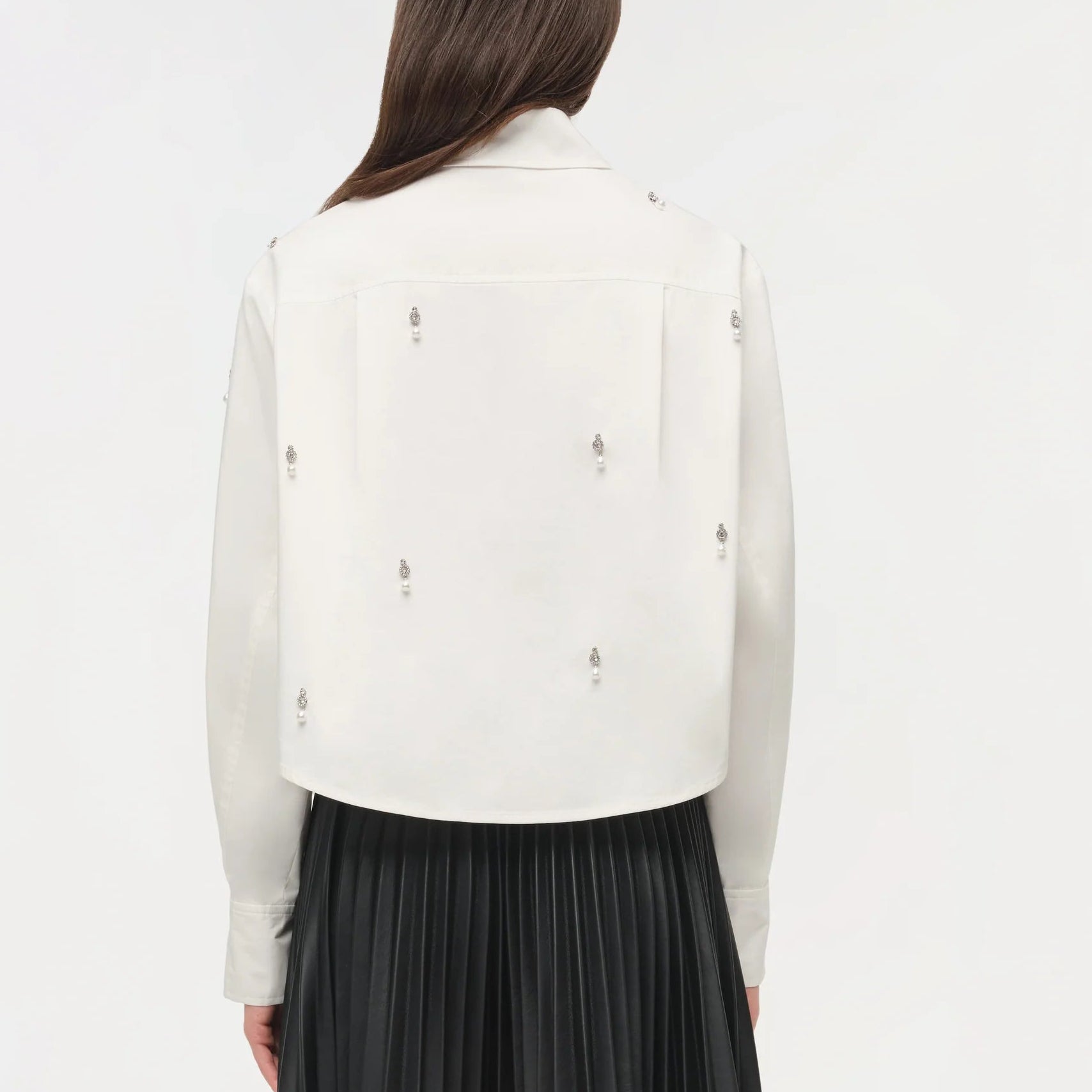 A person with long brown hair is seen from the back wearing the Simkhai Renata Embellished Cropped Shirt—white cotton poplin with crystal pearl details—and a black pleated skirt, set against a plain background.