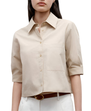 A woman wears the TWP Short Lived Top, a beige, short-sleeve cotton poplin button-up with a chest pocket and boxy fit, tucked into white pants and styled with a brown belt.