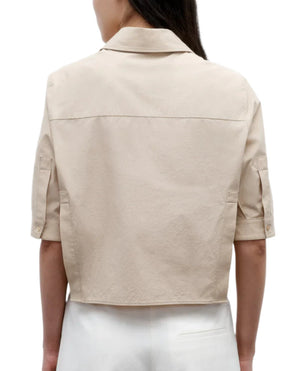 A person wearing the TWP Short Lived Top—a light beige, boxy fit short-sleeve collared shirt with buttoned cuffs—viewed from the back and paired with white pants.