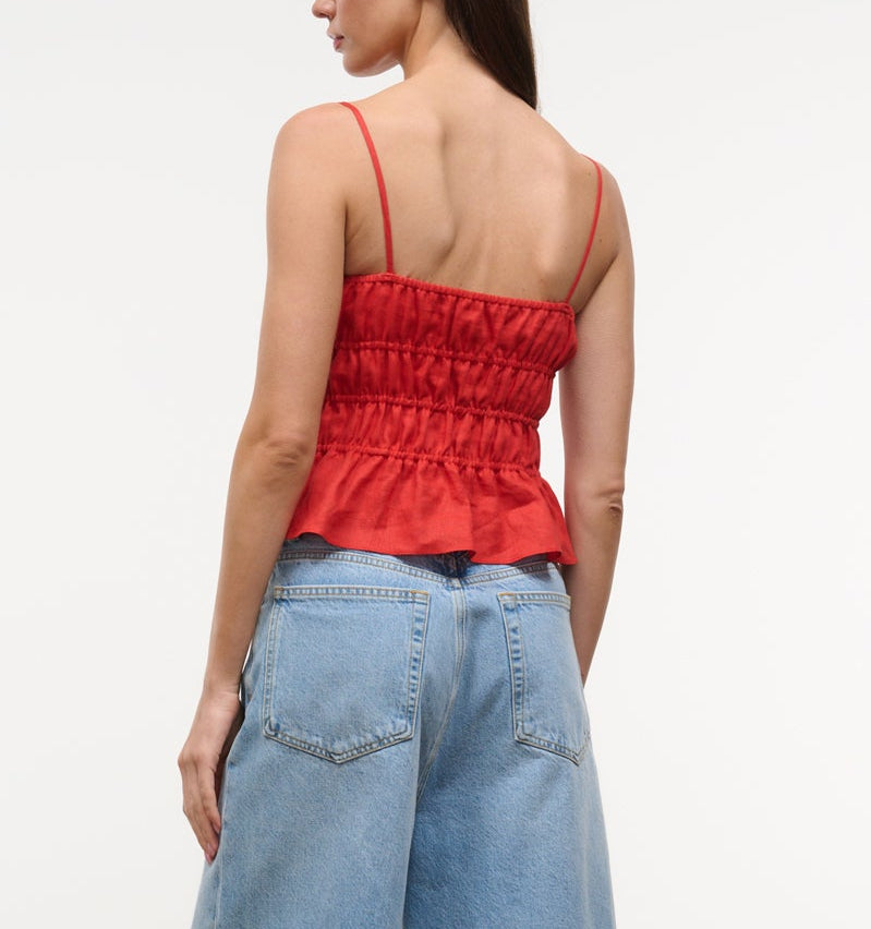A woman seen from the back wears the Staud Ayla Linen Tank, a red cami with thin straps and a smocked bodice, paired with blue high-waisted jeans.