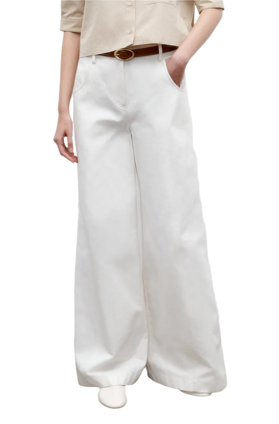 Model wears TWP’s Dee's Linen Trouser—wide-leg, white cotton linen—paired with a beige shirt, brown belt, and white shoes, one hand in the pocket, set against a white backdrop.
