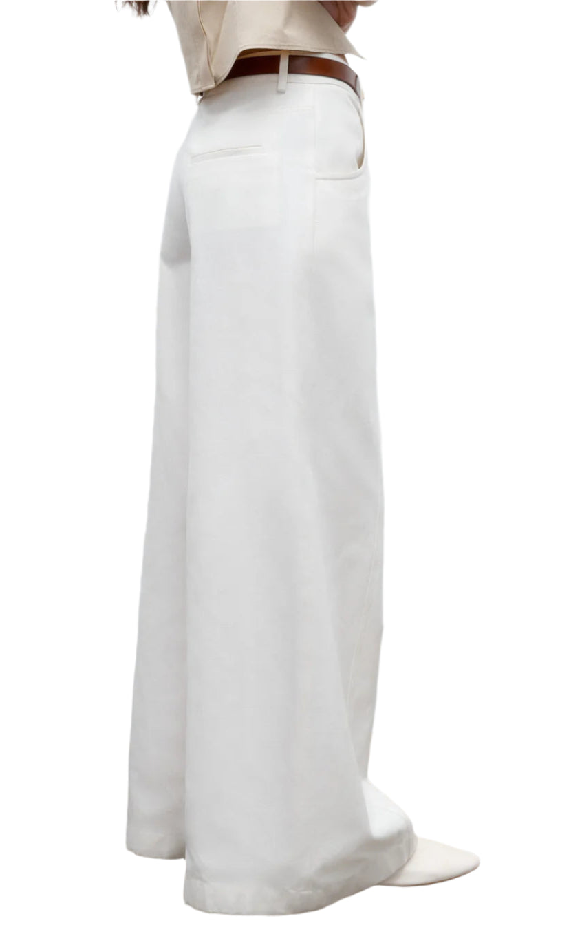 A person is shown from the side against a white background, wearing TWP's Dee's Linen Trouser—high-waisted, wide-leg cotton-linen pants—styled with a brown belt and white pointed shoes.