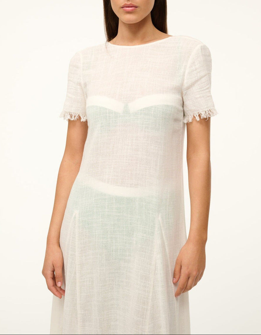 A woman wears the Staud Le Havre Dress, a sheer white cotton gauze short-sleeve style with frayed edges and minimal design, perfect as a swim coverup. Photo is cropped at shoulders and mid-thigh, revealing an undergarment underneath.
