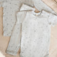 Two light grey baby outfits with a celestial print are displayed on a beige quilt: one is a long-sleeved shirt with pants, and the other is the Quincy Mae Baby Bamboo Short Sleeve Bodysuit.