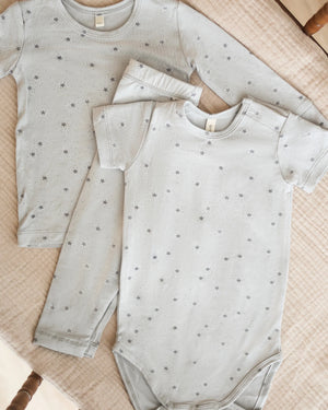 Two light grey baby outfits with a celestial print are displayed on a beige quilt: one is a long-sleeved shirt with pants, and the other is the Quincy Mae Baby Bamboo Short Sleeve Bodysuit.