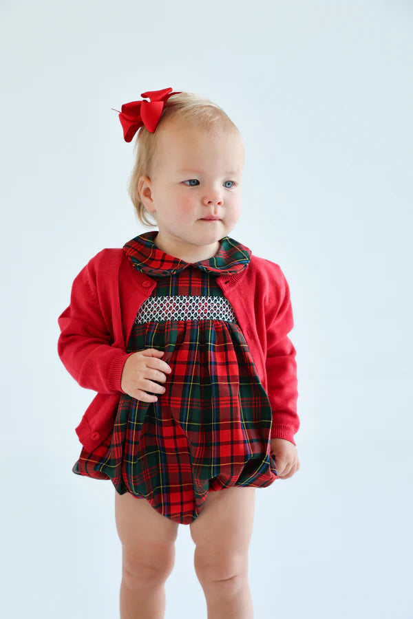 A toddler in The Beaufort Bonnet Company's Baby Girls' Dottie Hart Bubble, styled with a red cardigan and matching red bow, stands against a plain light background.