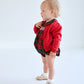 A toddler in The Beaufort Bonnet Company's Baby Girls' Dottie Hart Bubble stands against a plain white background, looking to the side.