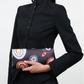 A woman in a black buttoned blazer holds the Staud Muse Beaded Clutch, featuring colorful circular patterns and a compact silhouette, against a plain background.