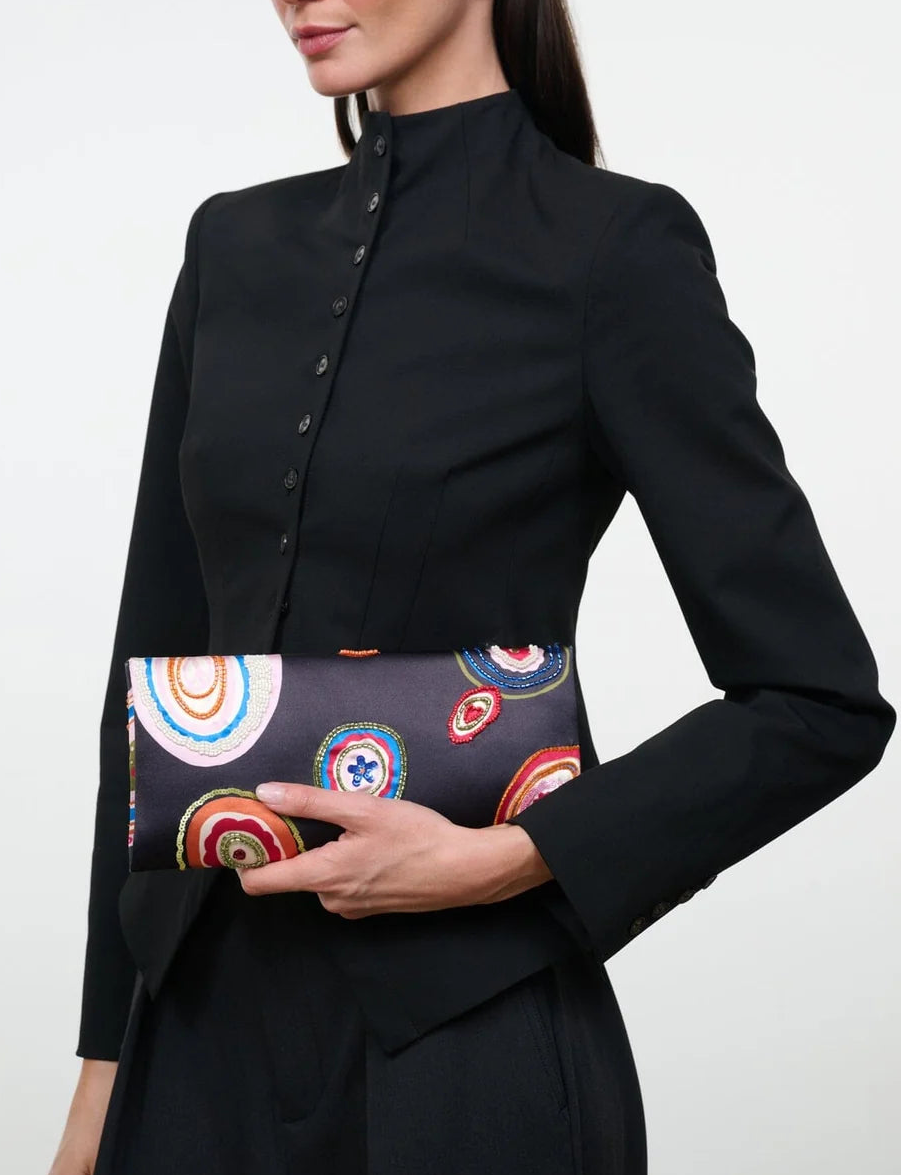 A woman in a black buttoned blazer holds the Staud Muse Beaded Clutch, featuring colorful circular patterns and a compact silhouette, against a plain background.