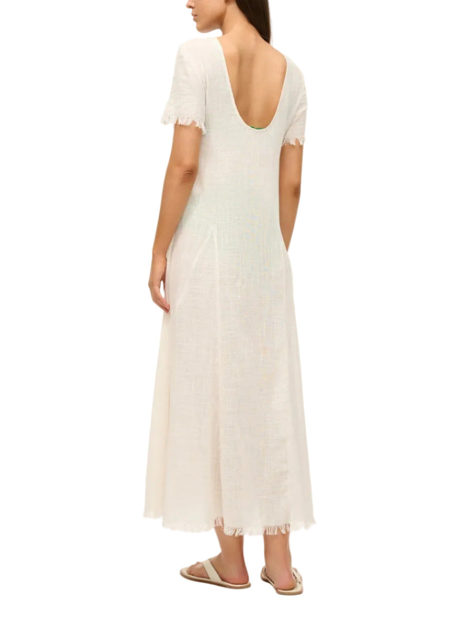 A woman wearing the Staud Le Havre Dress—a short-sleeve, off-white, ankle-length cotton gauze dress with a scoop back—stands facing away from the camera. Ideal as a lightweight beach swim coverup.