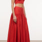 A woman stands against a plain background wearing the Staud Procida Linen Skirt, a high-waisted maxi skirt with a curved yoke, paired with a sleeveless red crop top and black sandals.