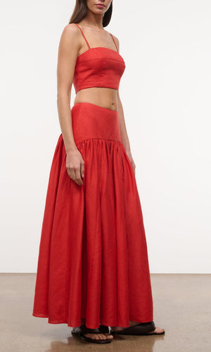 A woman stands against a plain background wearing the Staud Procida Linen Skirt, a high-waisted maxi skirt with a curved yoke, paired with a sleeveless red crop top and black sandals.