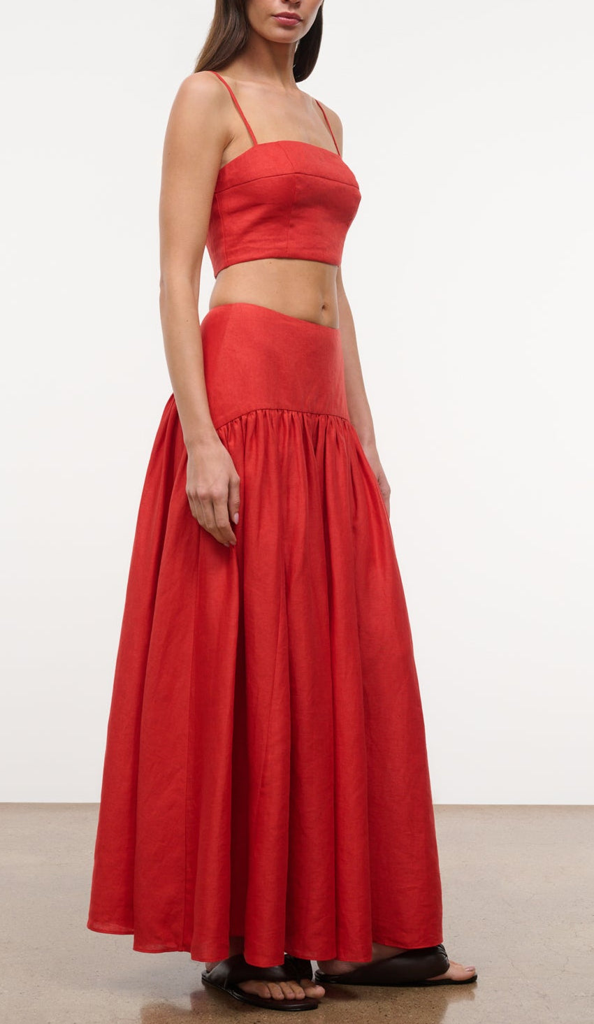 A woman wears the Staud Federico Linen Tank, a sleeveless red cropped top with adjustable straps, paired with a matching high-waisted, floor-length skirt, standing against a plain background.