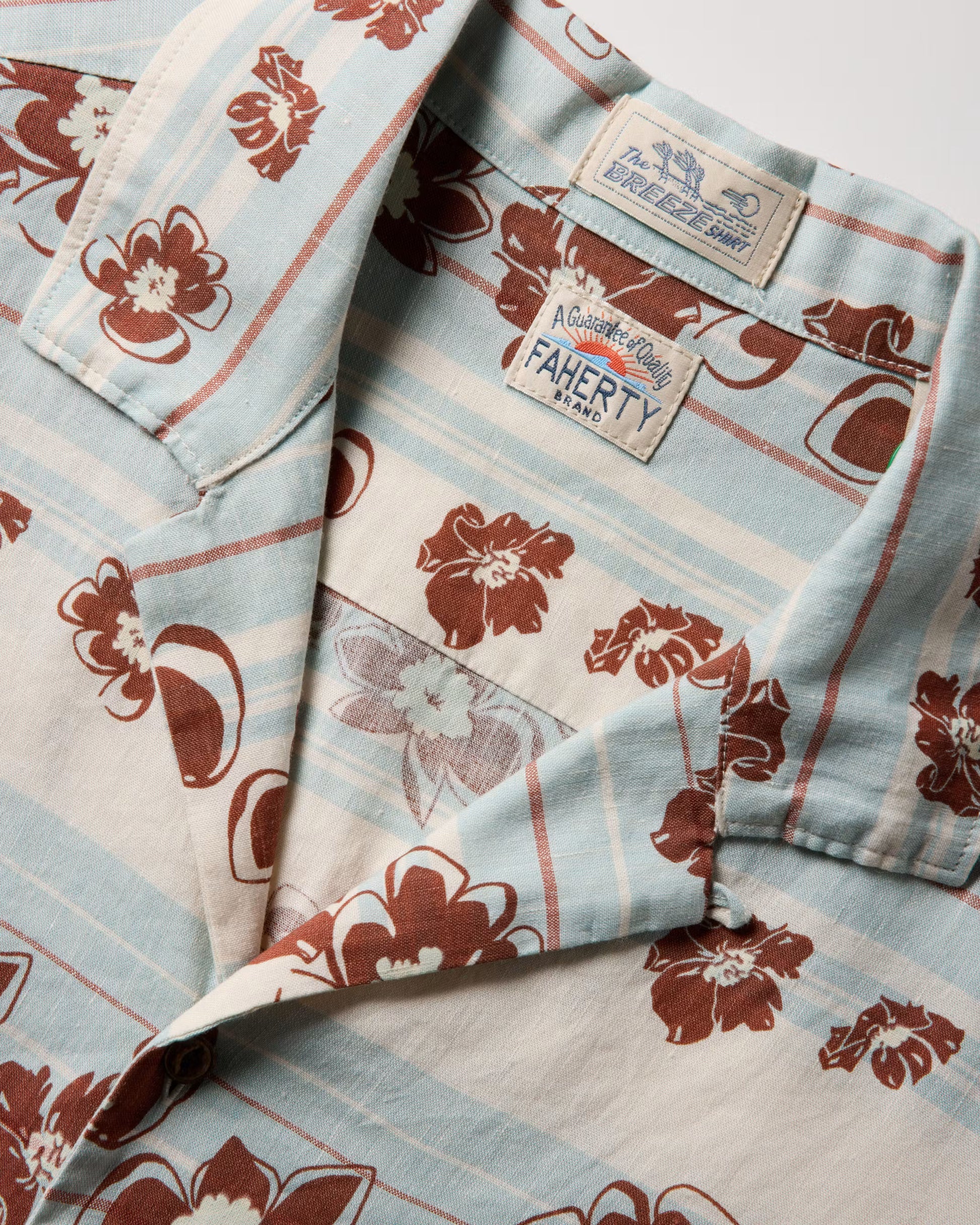 A close-up of the Faherty Breeze Camp Shirt, a light blue and cream button-up featuring brown floral patterns and two visible brand labels on the collar.